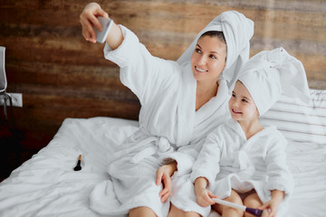 attractive woman and little child wearing white bathrobe and towel lying on bed taking selfie together after shower