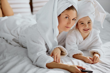 portrait of smiling caucasian woman and her little child girl dressed in bathrobe and towel looking at camera and holding tv remote