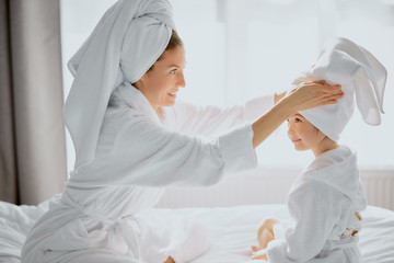 caucasian beautiful woman straightens towel to lovely daughter after shower, sit together on bed