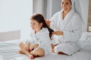 Fototapeta premium careful caucasian woman combing hair to awesome daughterdressed in bathrobe and towel after shower