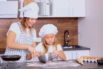 charming kid girl mixing flour in bowl with mother in kitchen, wearing apron and caps for cooking, family teamwork