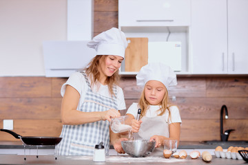 young caucasian blond woman sprinkle flour in bowl while her daughter mix it. Kid and woman wearing aprons isolated in kitchen