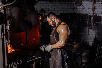 young strong muscular blacksmith wearing black apron and gloves for safety, stand preparing iron on fire, having powerful physique