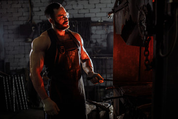 Fototapeta na wymiar young strong muscular blacksmith wearing black apron and gloves for safety, stand preparing iron on fire, having powerful physique