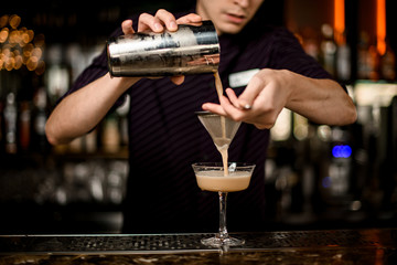 Male bartender pouring a alcoholic drink from the steel shaker to the cocktail glass through the sieve