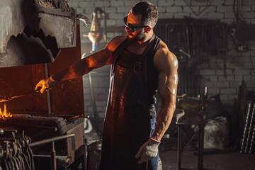 male blacksmith forging in his workshop, make iron products for manifacture of fireplaces and stoves. Use furnace for heating metal. Wearing protective eyeglasses and gloves