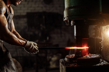 muscular forger man with beard using hydraulic press for steel arms manufacture at smithy, wearing leather apron or uniform