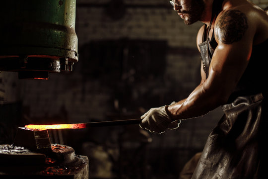 Young forger shaping metal on a jackhammer in the blacksmith workshop, wearing leather uniform. muscular body of man