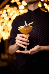 Bartender in the purple shirt serving yellow cocktail in the glass with a dried lemon and leaf in the clothespin