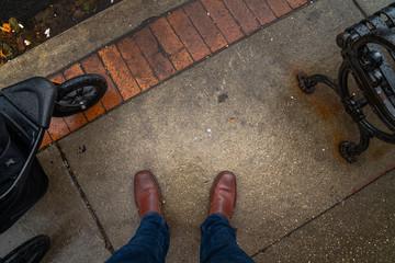 Looking down at the feet of a man wearing jeans and brown leather shoes standing on the concrete...