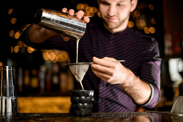 Male bartender pouring a alcoholic drink from the steel shaker to the black cocktail glass through the sieve