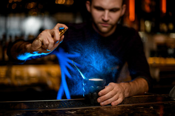 Male bartender spaying on a black glass with a delicious cocktail with essence under blue light