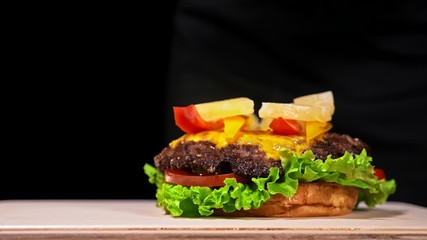 Craft burger is cooking on black background in black food gloves. Consist: red sauce salsa, lettuce, red onion, pickle, cheese, chilli green pepper, air bun and marbled meat beef. Not made ideal