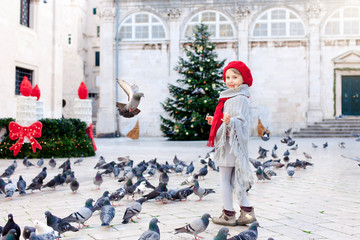 Christmas mood in Europe. Happy child girl feeds doves on square. Pigeons flying around cute kid....