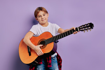 Fototapeta premium portrait of caucasian little boy 7-8 years old stand holding acoustic guitar, student of music school, posing with guitar. children interested in music and instruments. isolated purple background