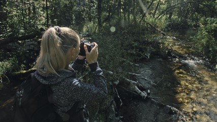 Traveler photographing scenic view in forest river. Wood bridge fallen tree. One caucasian woman shooting nice magic look. Girl take photo video on camera. Photographer travel with backpack. Outdoor.