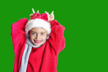 7 yaers old smiling girl with changing tees smiling in santa hat, red pullover, gray scarf, imitation of christmas dear with smile on green background. Natural light. Space for text