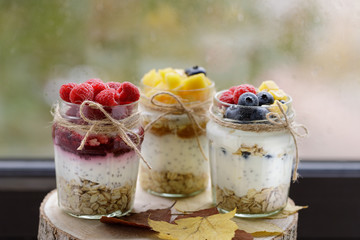 Healthy food, diet.  Glass jars of yogurt сhia pudding with oats, raspberries, mango slices, blueberries and jam on a wooden tray. Close up, with copy space