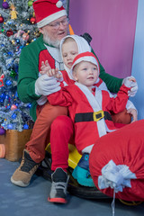 Happy children with dad are dressed in festive costume of Santa Claus and snowman, carrying decorated Christmas tree with balls and garlands on yellow car, accompanied by rocking horse. Christmas Eve