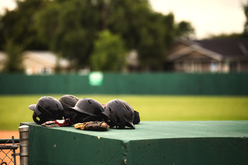 Closeup of four black baseball helmets and a glove on top of the dugout with a blurred baseball diamond in the background in summer - obrazy, fototapety, plakaty