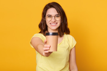 Studio shot of happy Caucasian woman holds take away cup of coffee or tea, having joyful facial expression, enjoying hot beverage, has coffee break, posing isolated over yellow studio background.