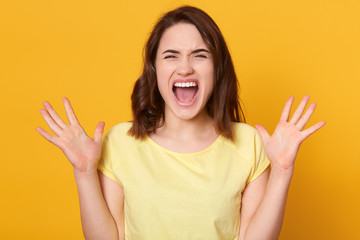 Portrait of expressive angry woman posing with closed eyes and screaming in studio isolated over yellow background, girl keeping hands up, female with dark hair, being in bad mood, being anger.