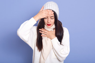 Image of upset attractive young girl closing her eyes, touching her forehead and neck, feeling unwell, having cold, running high temperature, wearing white hat, sweater and scarf. Health concept.