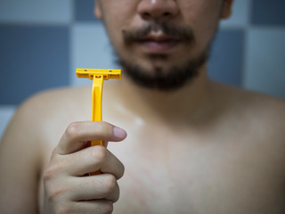 Selective focus on hand holding yellow Shaver with blur man has messy beard and mustache in bathroom background - 306732723