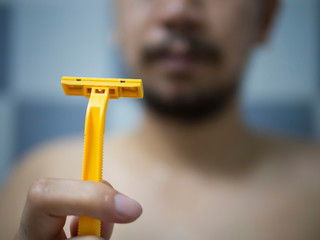 Selective focus on hand holding yellow Shaver with blur man has messy beard and mustache in bathroom background - 306732737