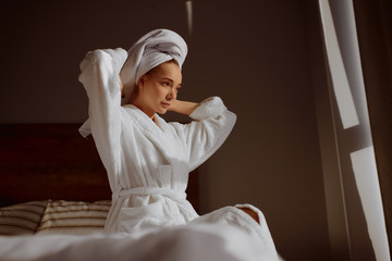 Charming beautiful female sitting on bed in bedroom, looking away with confidence, expressing calmness, resting after shower, background of gray wall, indoor shot from below