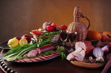 Sausage, ham, a bottle of red wine and glasses with wine surrounded by vegetables on the table.