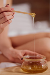 Photo of liquid sugar paste flowing down to glass jar from wooden stick, female preparing everything for depilation procedure, beauty and health treatment concept