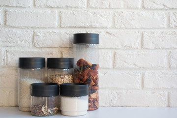 transparent jars with cereals, sugar, seasonings and dried fruits on the table on a brick background