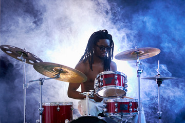 Fototapeta na wymiar black young guy with dreadlocks wearing eyeglasses sit beating on drums with sticks, with naked skin isolated over smoky background