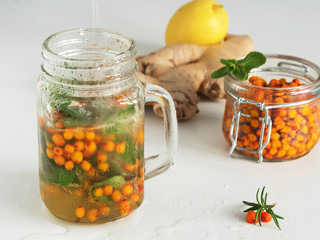 Hot drink in a container with a handle on a white background, next to the ingredients for tea. Winter drinks. Tea with mint, ginger, lemon, sea buckthorn and honey. Close-up.