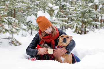 Fototapeta na wymiar Beautiful smiling woman is sitting on a snow with her two dogs wrapped in red checkered plaid and two cup of hot tea in winter coniferous forest.
