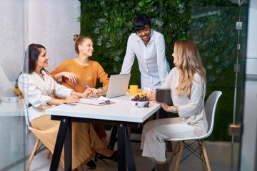 Confident young african lady in white shirt stand talking to caucasian young women, teaching business mentoring, discuss strategy in modern office