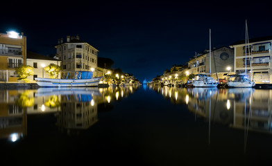 Fototapeta na wymiar Nighttime shot of Port of Grado in Italy mirroring in water, with passageway to adriatic sea and thawed boats