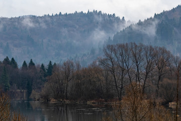 Fototapeta premium A small river at the foot of the mountains. The tops of high mountains are hidden by clouds. Fog. Autumn landscape. Panoramic view.
