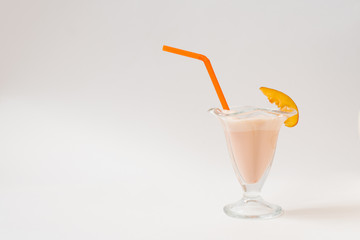 A glass jar with an orange plastic tube and a beautiful pale orange milkshake in it. A glass with a piece of nectarine on a white background.