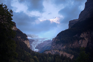 Scenic view of the alpine glacier in dusk. Snowy peaks emerge from the clouds. Summer alpine landscape in twilight. Grindelwald area. Switzerland.