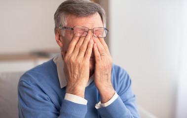 Senior Man Massaging Tired Eyes Sitting On Couch At Home