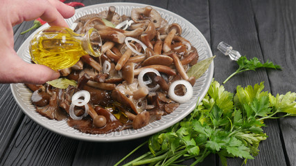 Pickled mushrooms in a large plate and fresh green vegetables on a black wooden table, a female hand pours oil from a bottle