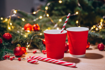 Obraz na płótnie Canvas Red paper cup on christmas background with bokeh. Red Christmas balls, fir branches, a garland with bulbs lie on the table.