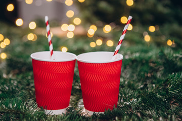 Red paper cup on christmas background with bokeh. Red Christmas balls, fir branches, a garland with bulbs lie on the table.