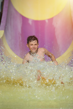 Happy smiling boy gliding down colorful water slide in water park during his summer holidays. He is making numerous splashes around.