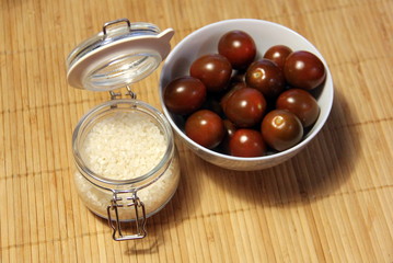 Small dark tomatoes in a white bowl and a glass jar with rice on a table