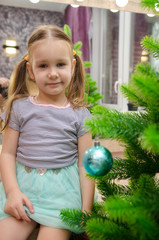 Fototapeta premium Adorable little girl decorating a Christmas tree with colorful glass baubles at home.Child decorating Christmas tree at home. Kids decorate living room and fireplace for Christmas.
