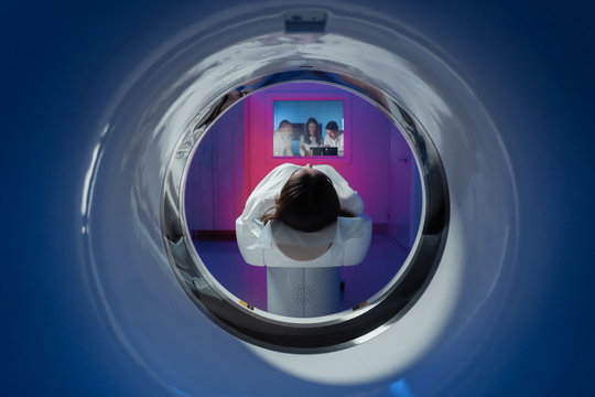 The girl patient is lying in the tomograph and waiting for a scan. Three doctors from the exam room look at the pictures