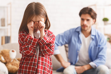 Little girl crying after arguing wIth dad, angry father behind
